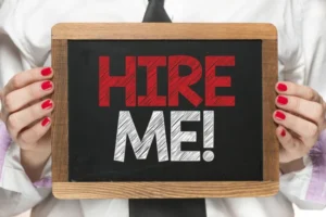 Woman with bright red fingernails holding Hire Me sign on a small blackboard with wood frame. Hire is in red chalk, and Me is in white chalk.