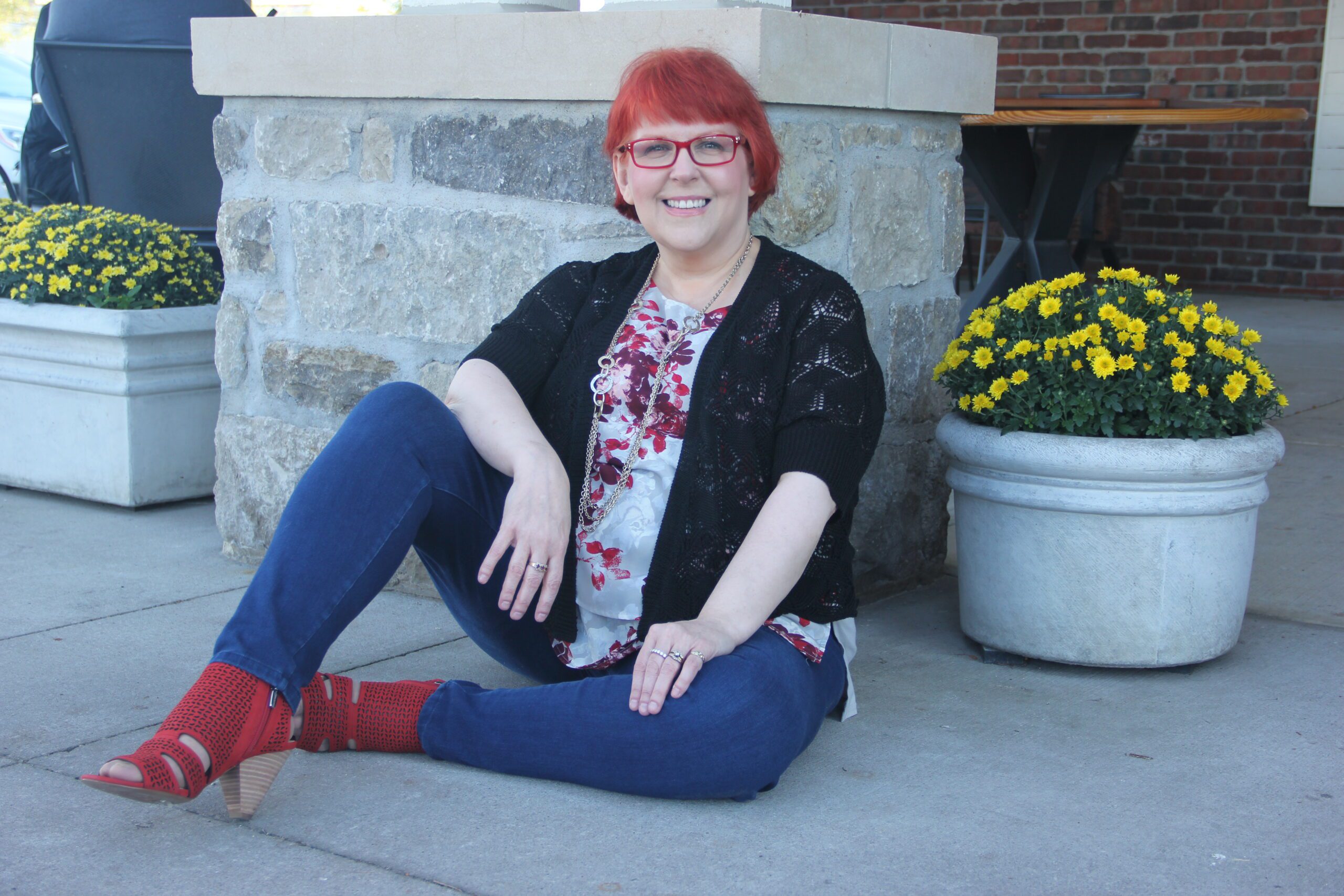 Karen Silins seated on ground in front of pillar and next to a pot of flowers, wearing black sweater shrug, floral blouse, blue jeans, and bright red heels.
