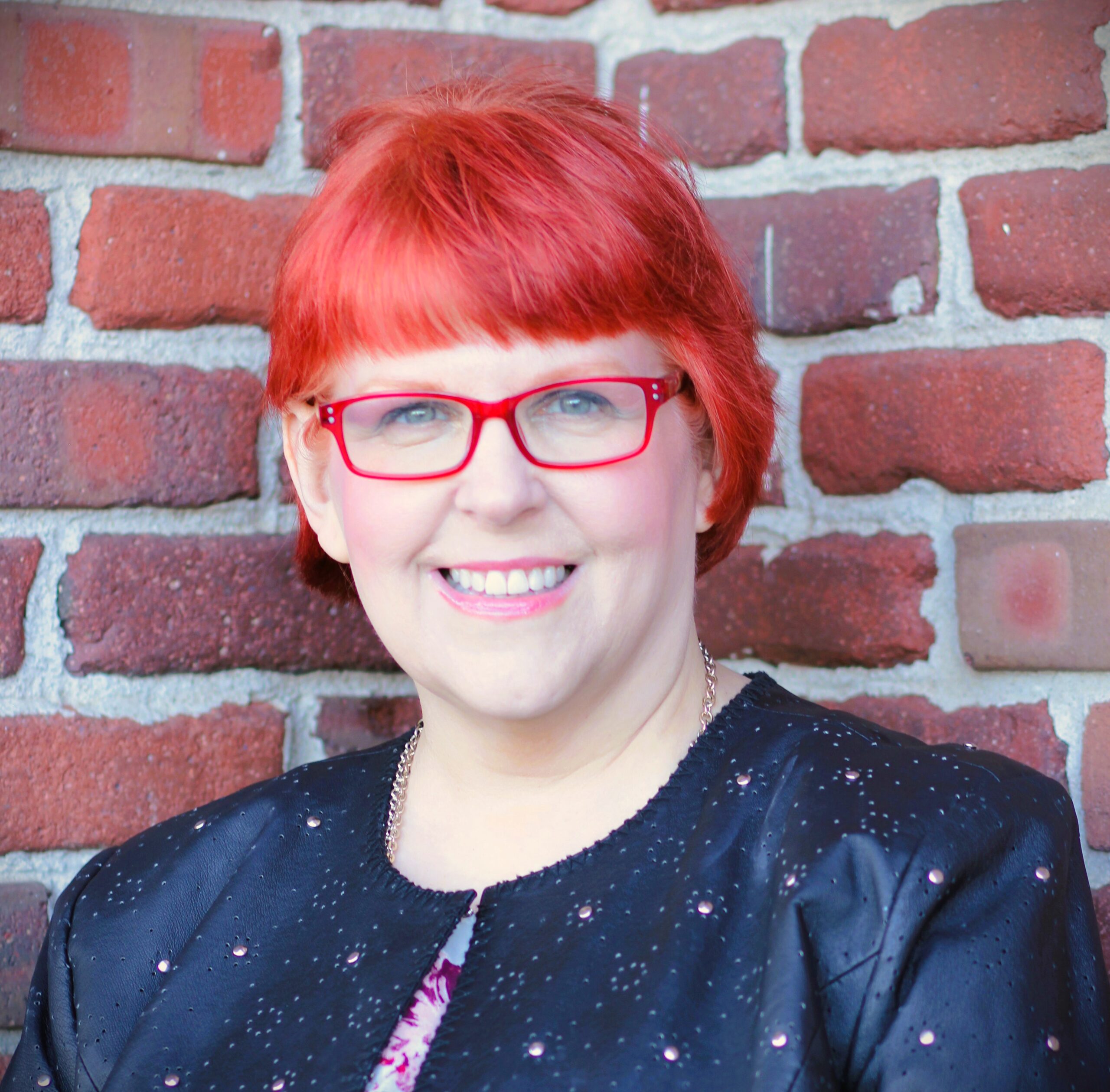 Karen Silins standing in front of a brick wall, smiling, with red glasses on, black stylish jacket, and bright red hair.