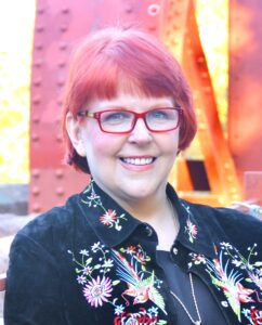 Karen Silins in front of bridge, wearing black jacket with floral embroidery, red glasses.